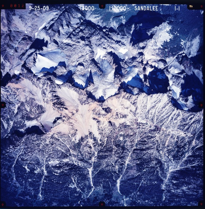 Color aerial photograph of glacier and snowfields on mountain peak with forested valleys below.