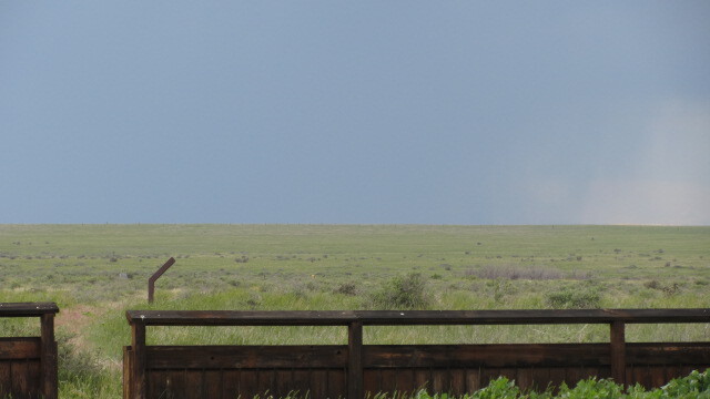 A view of a plain landscape with a dark sky and rain in the distance, behind a metal barrier.