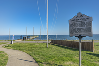 An interpretive sign stands on green grass, with a pier reaching out over the horizonless waters beyond. 
