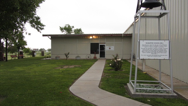 The front entrance of a beige building with a sign, two potted plants, and a concrete walkway.