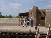 People are standing near an adobe structure under a sunny sky.