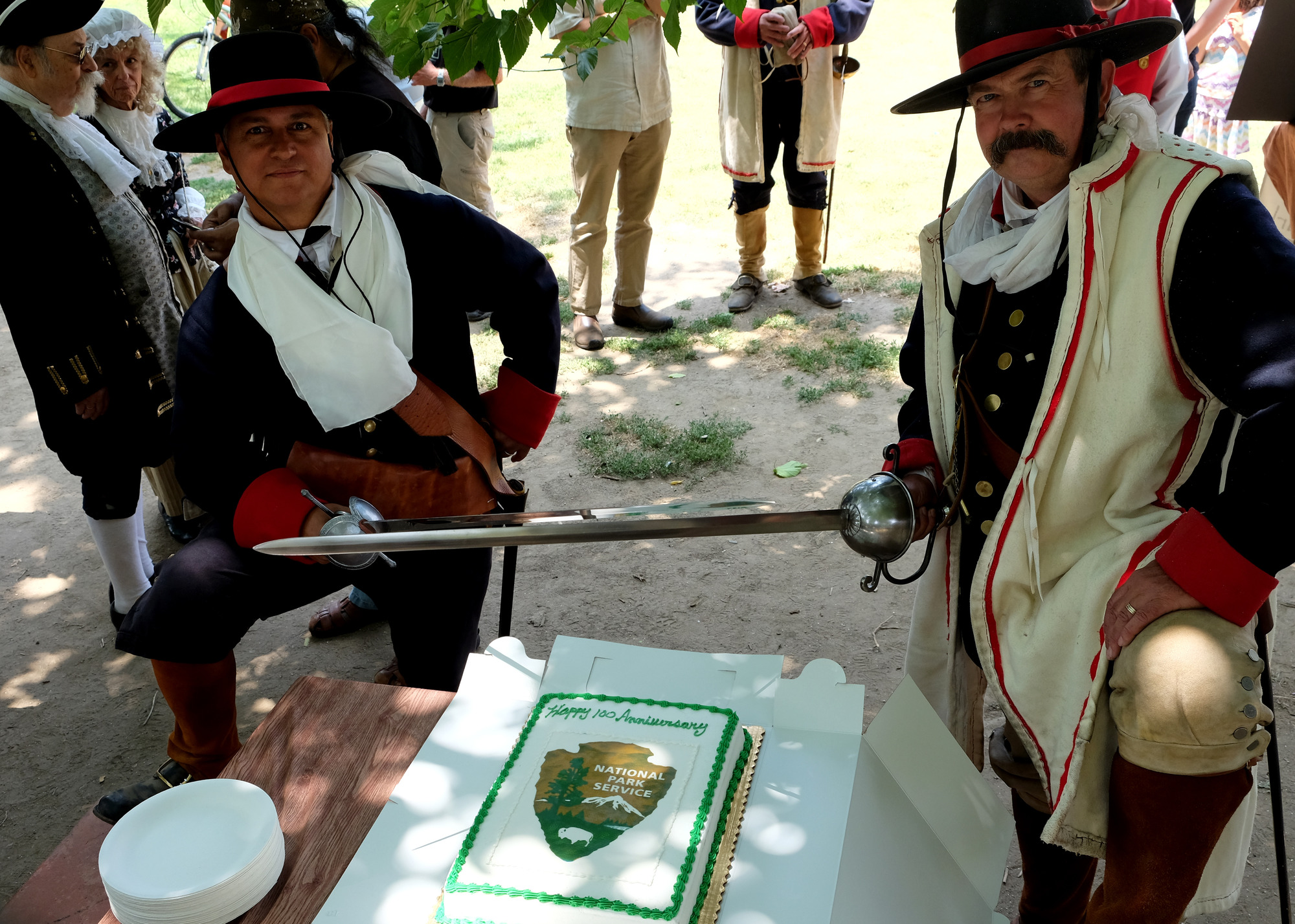 Two volunteer living historians dressed as soldados prepare to cut the National Park Service birthday cake with their sword. 