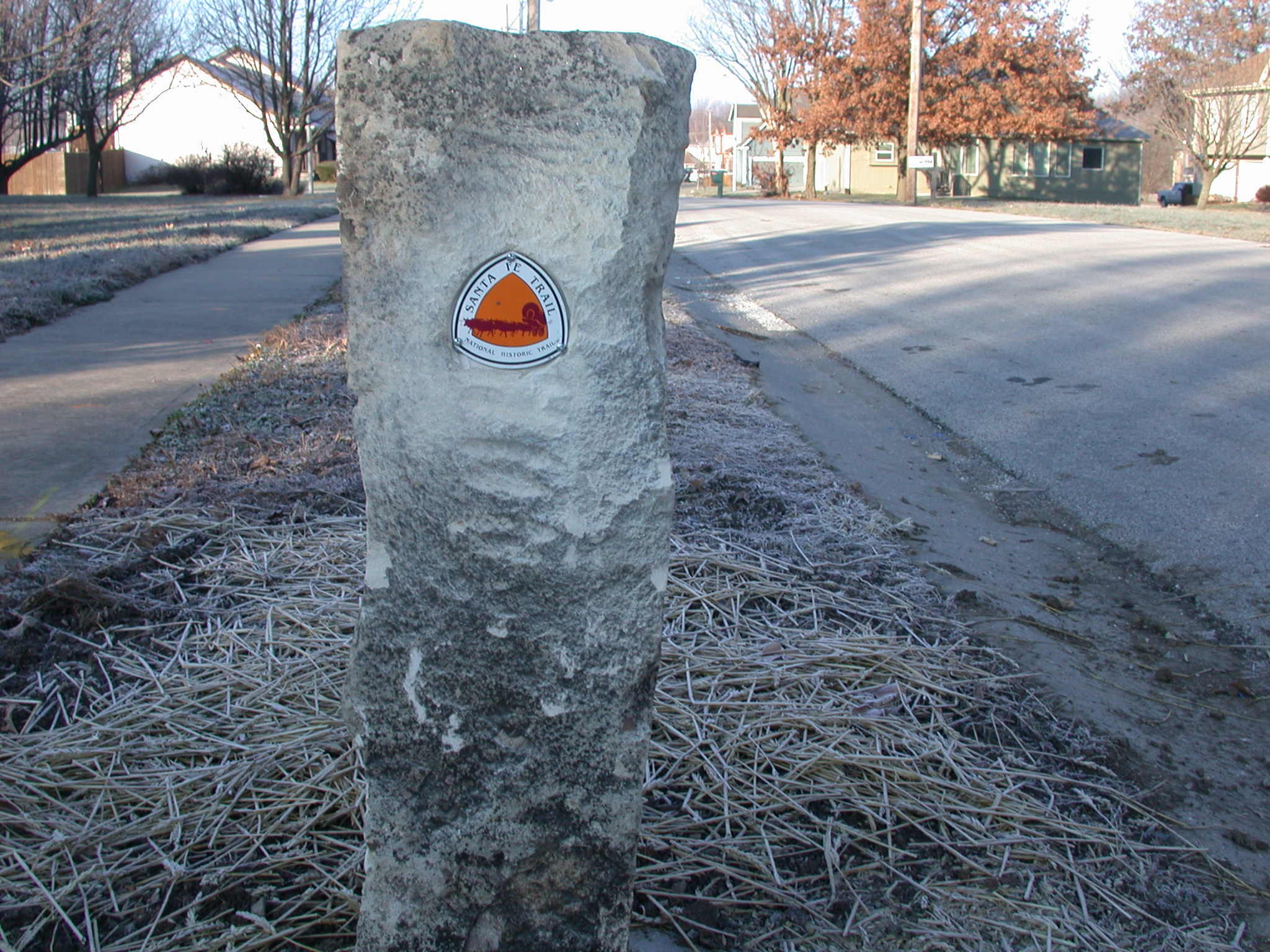 A trail marker on a concrete post beside a sidewalk, with houses and a street in the background.