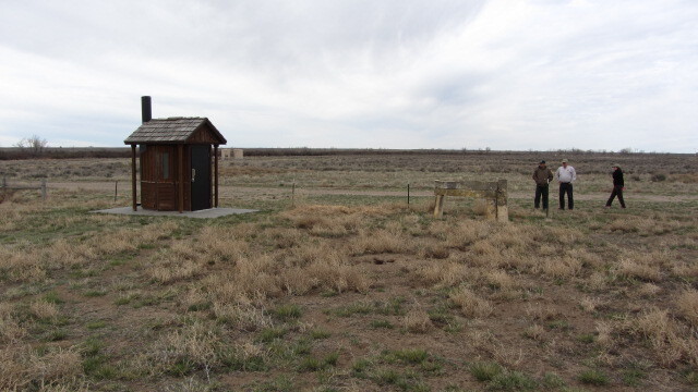 A group of people next to a wooden outhouse surrounded by a grassy field.