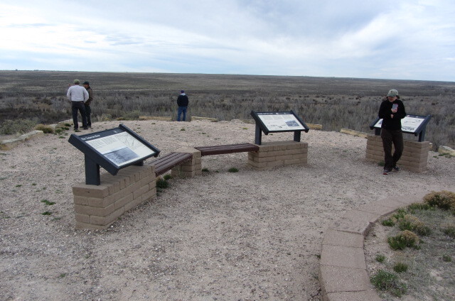 People are reading information on wayside panels at a viewing area overlooking a wide, open landscape.