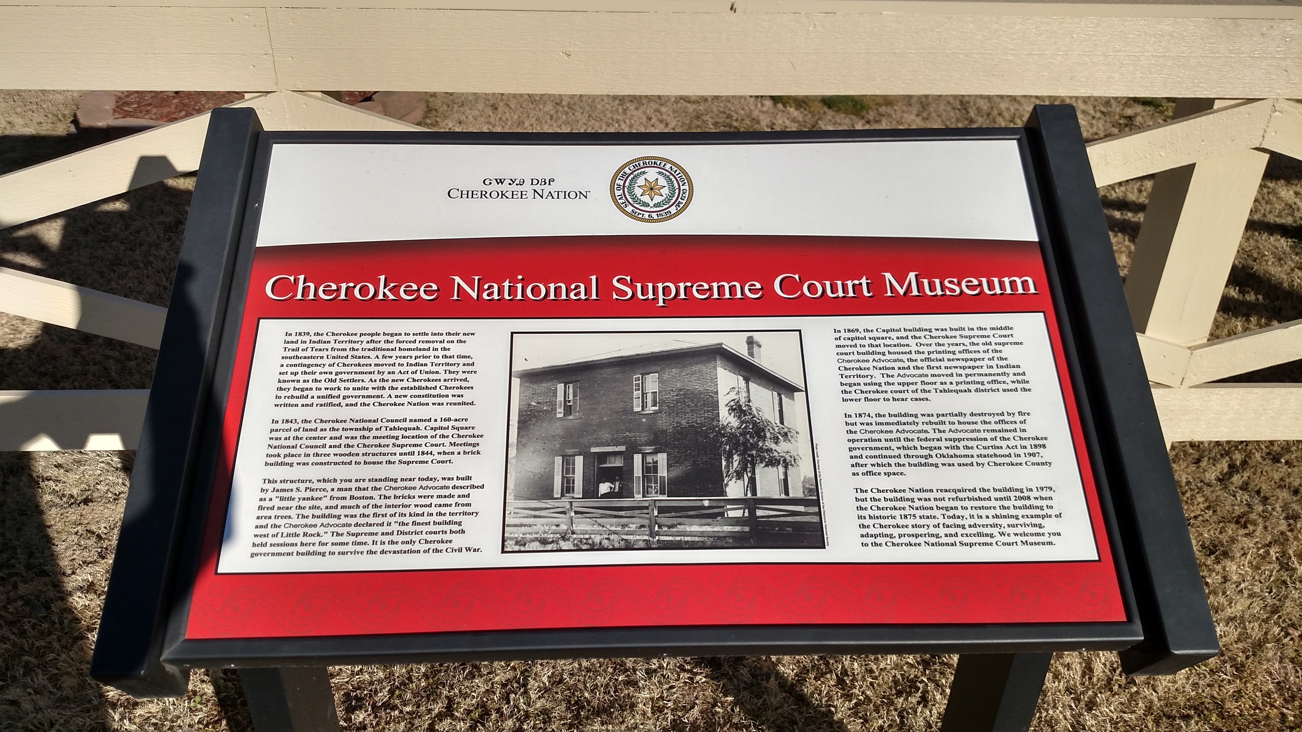 A wayside with text and a photograph, mounted on a stand with a bench in the background.