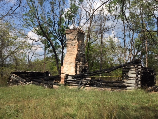 A burned out log cabin in the middle of a wooded area.