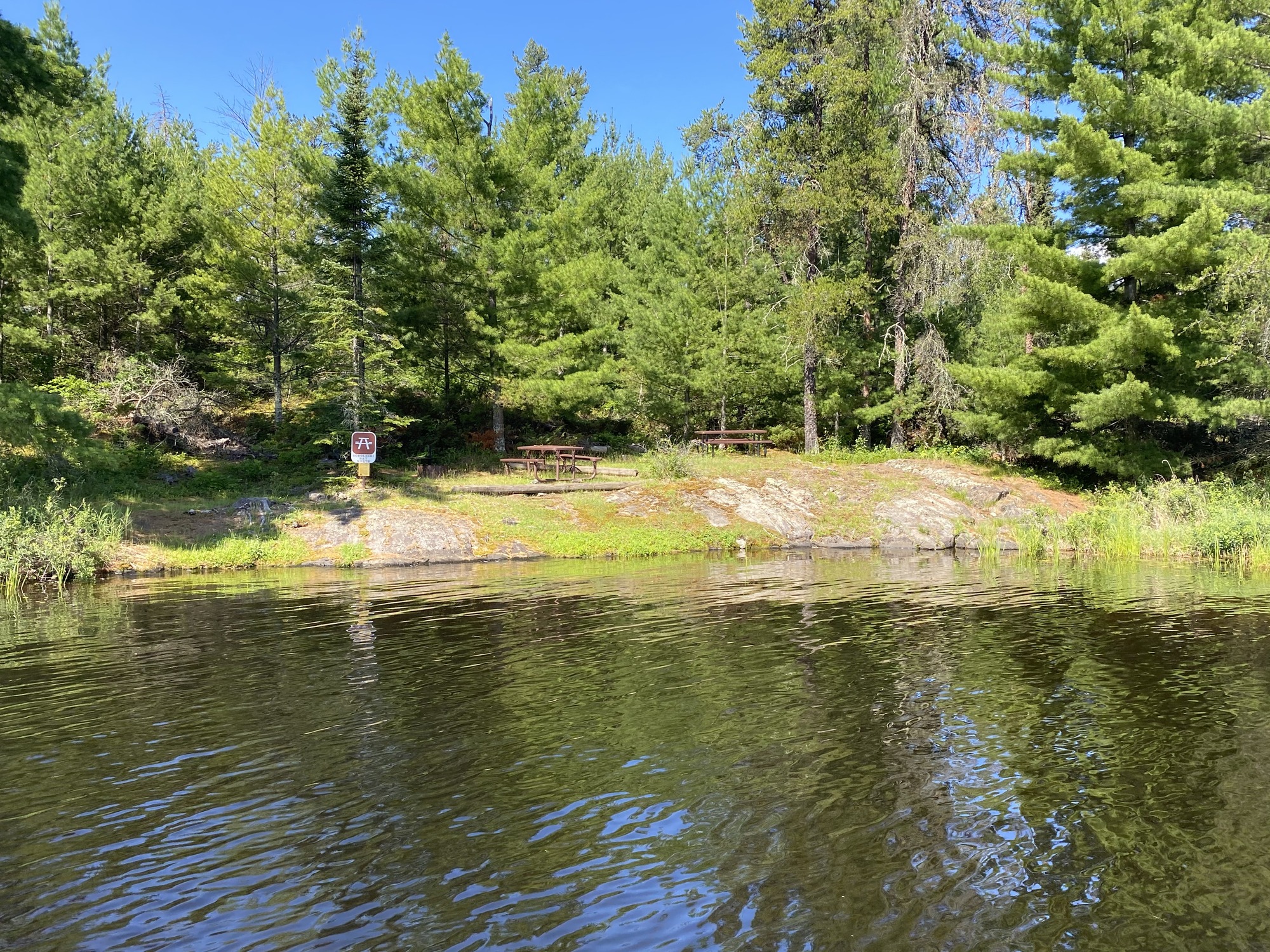 Tranquil lakeside scene with dark, reflective water and a rocky shore; a bench and sign nestled among tall green pines.