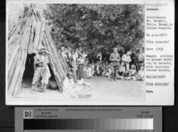 Mr. Harwell, Chris Brown in Native American regalia. Maggie sitting on ground working on baskets, with tourists behind museum.