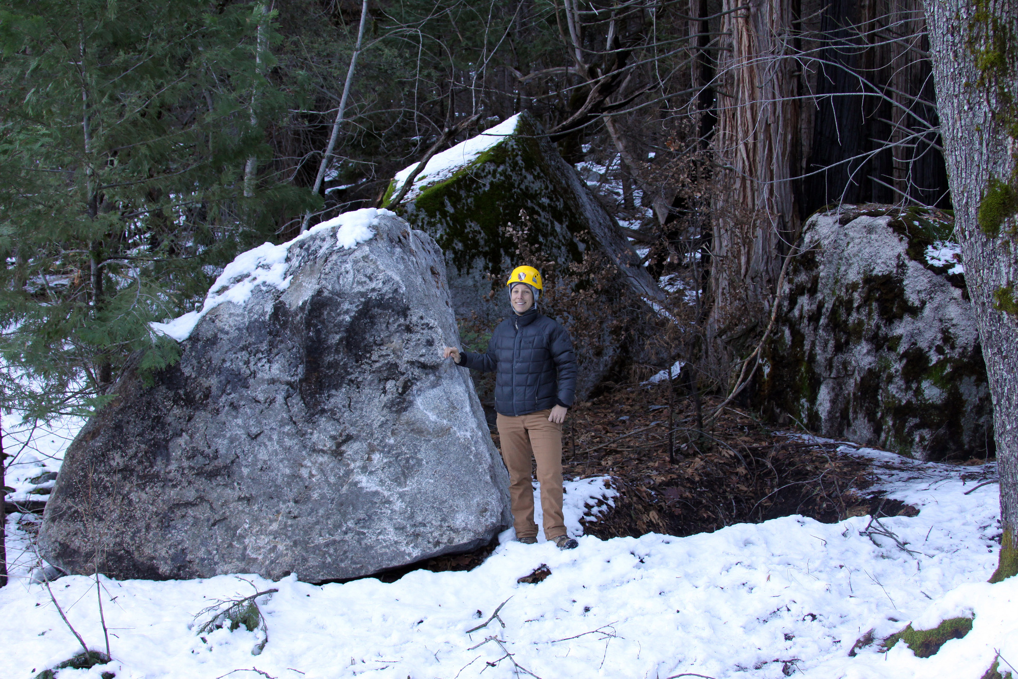 A person stands next to a large boulder in a forested, snow covered area.