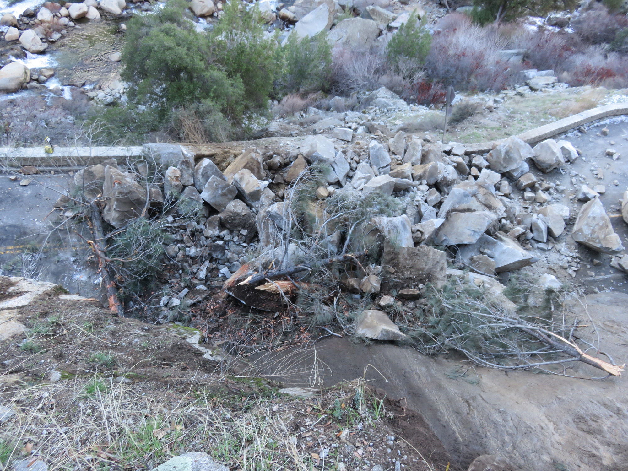 View from above looking down at fresh boulders and downed trees covering both lanes of a road and debris extending down the slope below.