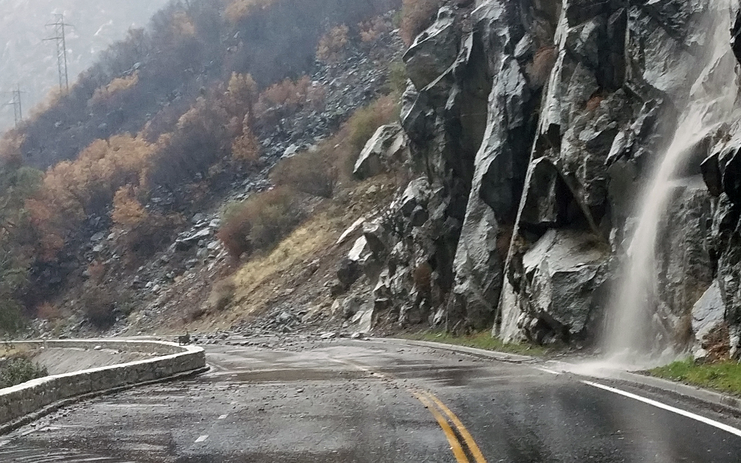 Water rushing down a rock slope and pooling on the road with rock debris, sand and silt across both lanes.