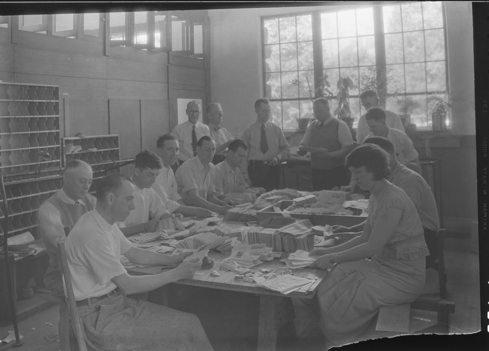 Postal Clerks at Yosemite Post Office for stamp issue 1st of the Nat. Park stamp series, the Yosemite 1 cent stamp. "It was necessary to supplement the regular Yosemite postal staff with ten extra clerks to handle the first day covers of the new National Park Service 1 cent stamp. Distinguishable in the background are (standing left to right): Postmaster F.C. Alexander (dark rimmed glasses) Mr. Charles F. Anderson, Post Office Dept., Washington (Profile), and Robert E. Fellers, Division of Stamps Supt. (silver belt buckle). On Monday, July 16, the first of the National Park stamp series, the Yosemite 1 cent stamp, with El Capitan Rock predominant, was put on sale; on that day, half as many one cent stamps as are ordinarily sold in Yosemite in a year were cancelled on letters. First day covers went all over the globe." From the July 1934 monthly Superintendent reports.