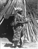 Chielf Le-me in front of Um-a-cha in back of Museum. [Original print in collection. Wearing mixture of traditional and non-traditional Miwok garb.]