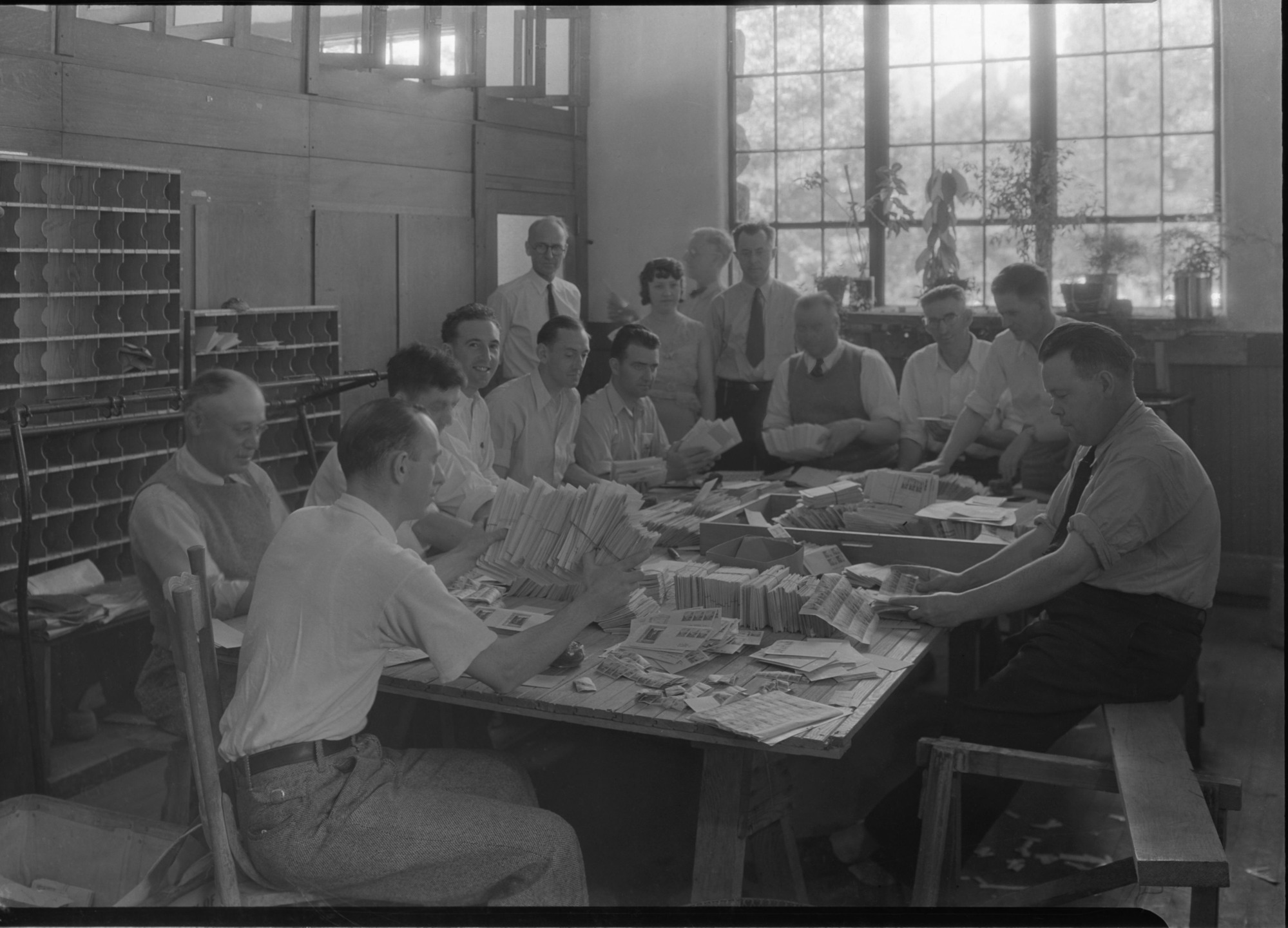 Postal Clerks at Yosemite Post Office for stamp issue 1st of the Nat. Park stamp series-the Yosemite 1 cent stamp. Distinguishable in the background are (standing up from left to right): Postmaster F.C. Alexander (dark rimmed glasses); Mr. Charles F. Anderson, Post Office Dept., Washington (profile); and Robert E. Fellers, Division of Stamps (silver belt buckle). See also July, 1934 monthly Superintendent Reports.