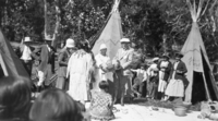 Alice Wilson in buckskin dress with back to camera. James Schwabacher wearing cap and holding large basket by Tina Charlie.