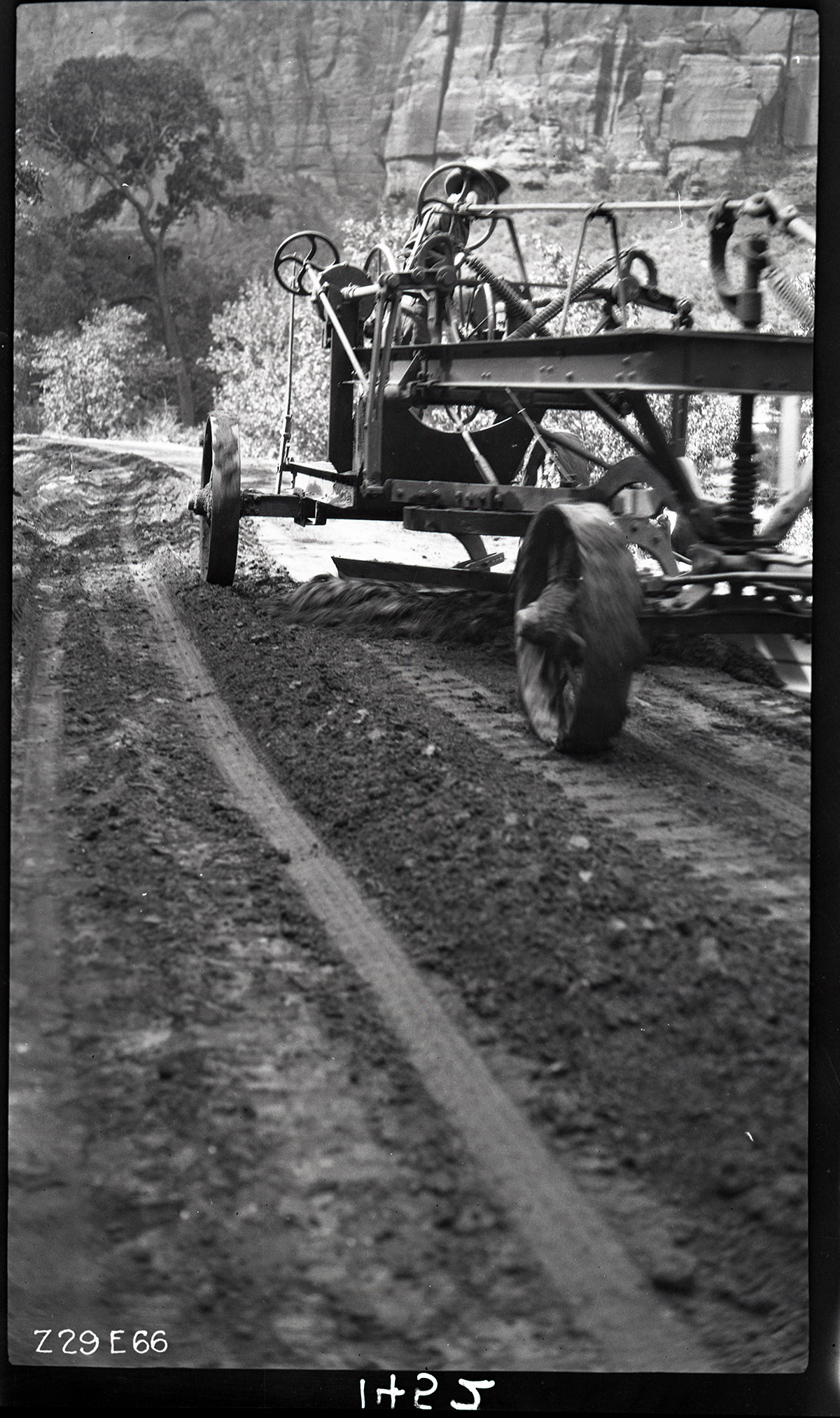 Worker driving a road grader in Zion Canyon. Labeled 'experimental road paving. Processing sand and bitumul 60'.