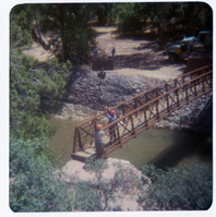 Two men guiding new Grotto footbridge into place as it is lowered on pulley system. Note bank revetments in background.