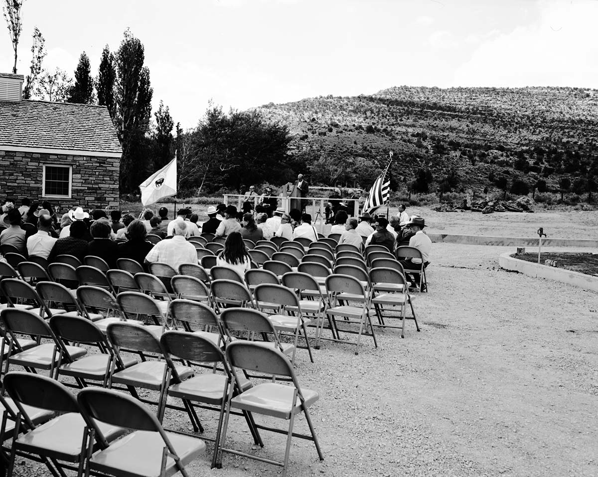 National Park Service Midwest Regional Director J. Leonard Volz addressing visitors at the dedication of new Tribal and National Park Service Visitor Center and 50th anniversary at Pipe Spring National Monument. United States and Department of the Interior flags at either side of grandstand.