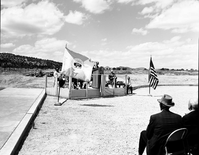National Park Service Midwest Regional Director J. Leonard Volz addressing visitors at the dedication of new Tribal and National Park Service Visitor Center and 50th anniversary at Pipe Spring National Monument. United States and Department of the Interior flags at either side of grandstand.
