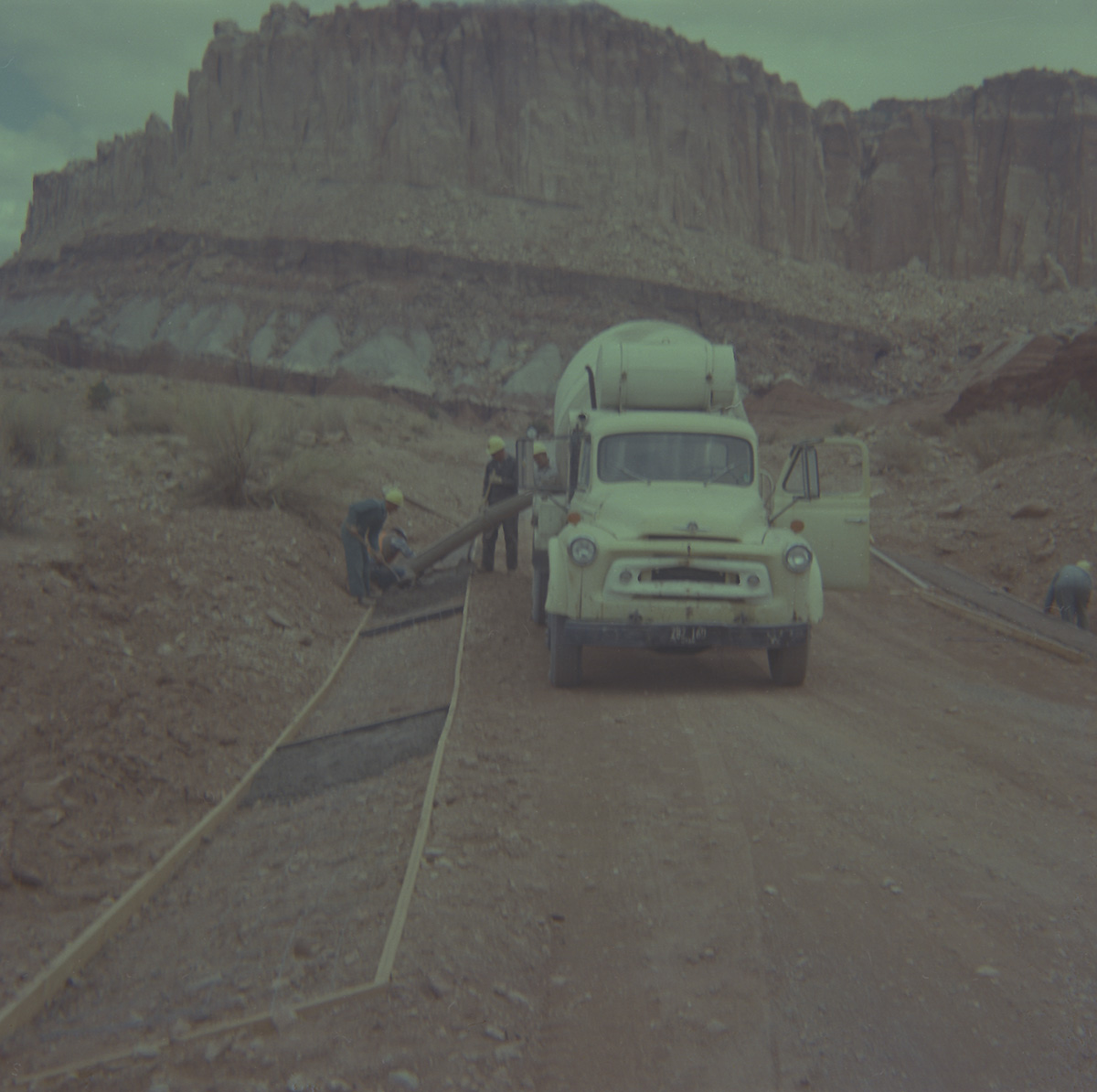 Cement truck laying cement while men watch on and assist in road repair.