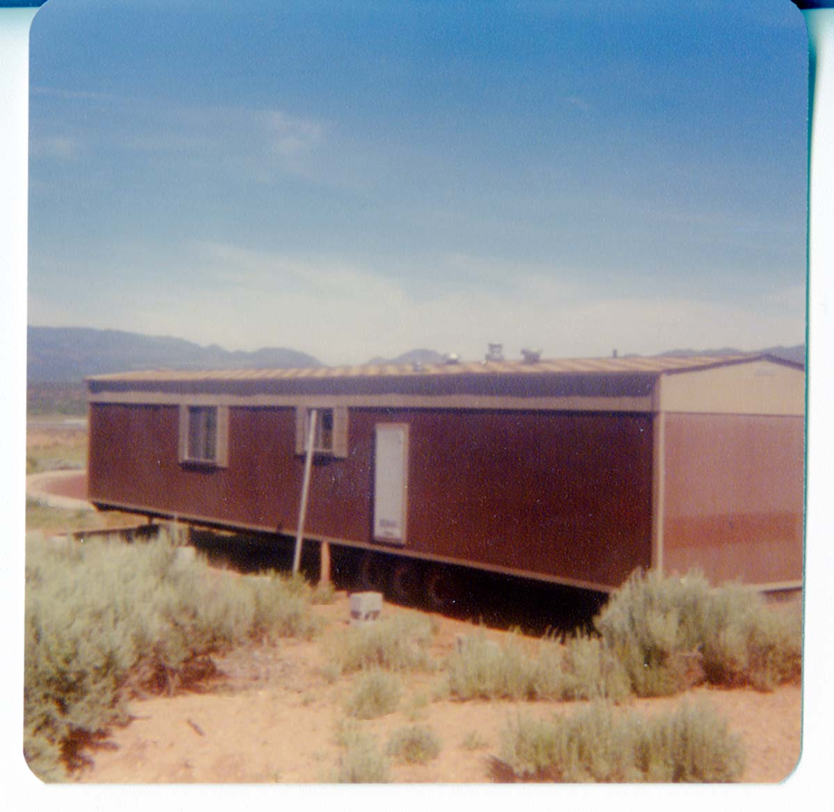 Single wide trailer home loaded onto truck trailer. Sage brush in foreground, mountains in background.