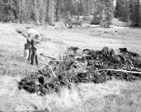 Two men watching water level in trench opened for water exploration, nearby large dirt pile. Water pump and gauge set up in trench.