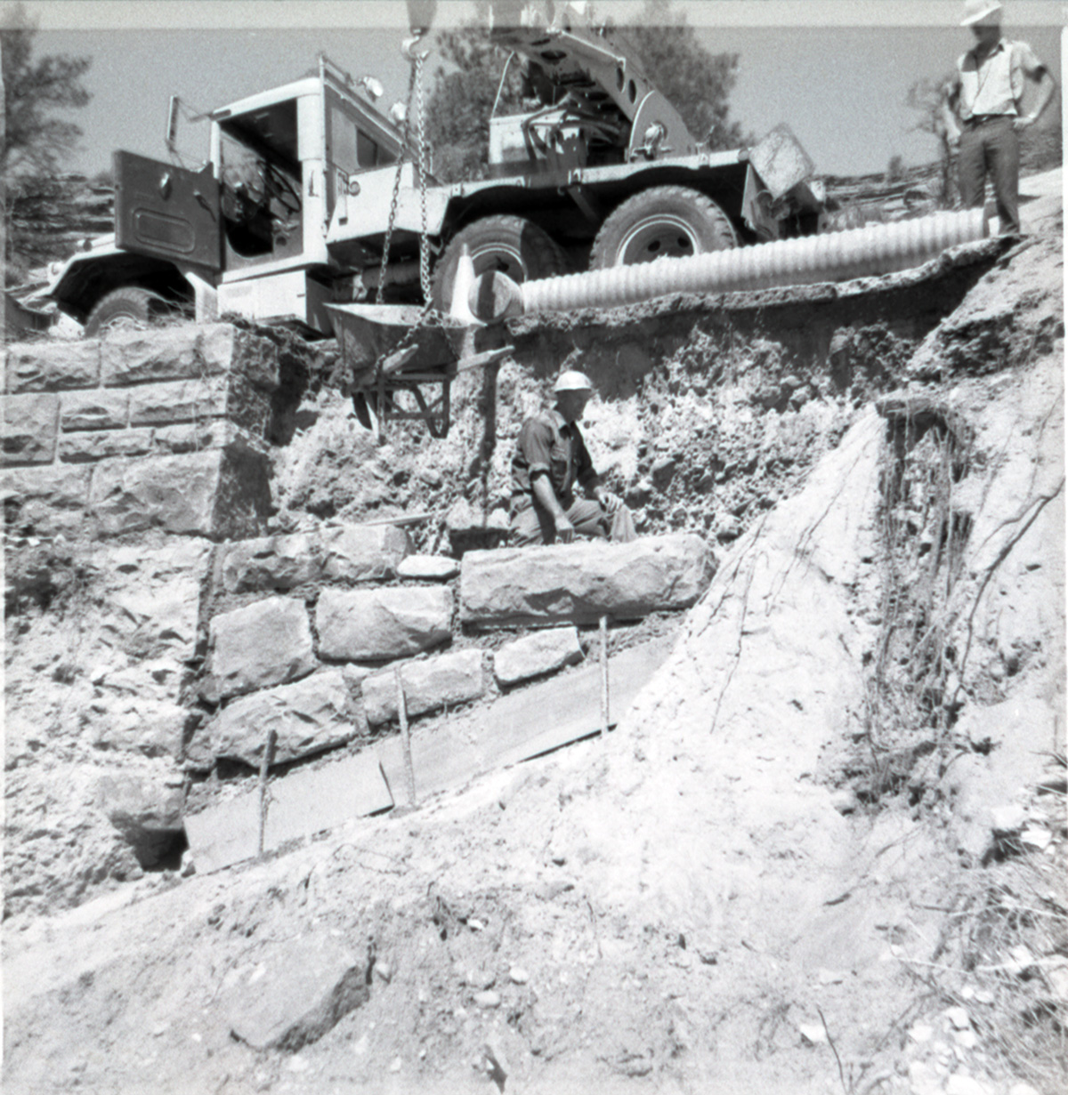 Men working on repairing retaining wall along East Rim road while crane lowers wheelbarrow full of building materials.