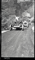 Worker driving a road grader following another worker on a caterpillar tractor on the floor of the valley road spreading paving materials. Labeled 'experimental road paving.'