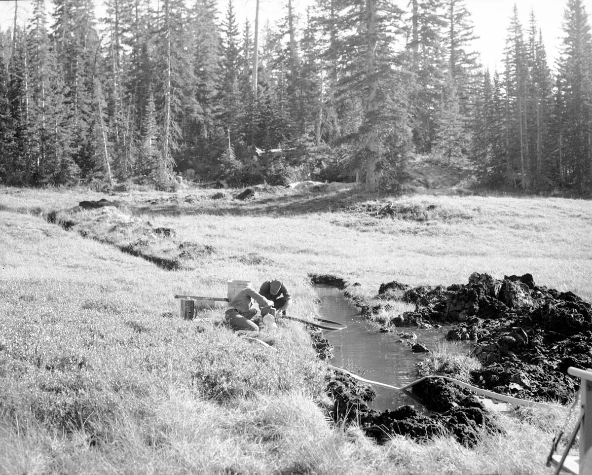 Two men adjusting pump next to overflowing trench opened for water exploration, nearby large dirt pile.