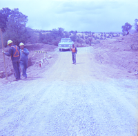 Men standing and discussing road construction and repairs with construction vehicle in background.