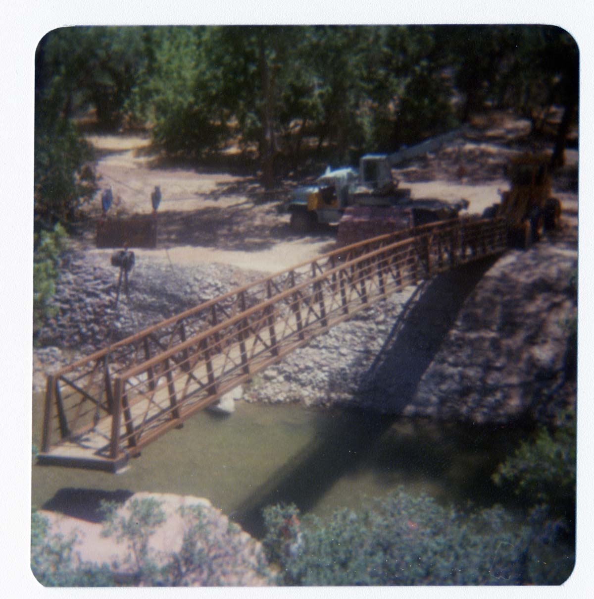New Grotto footbridge being lowered into place by pulley system, crane and backhoe anchoring one end of bridge. Note riverbank revetments.