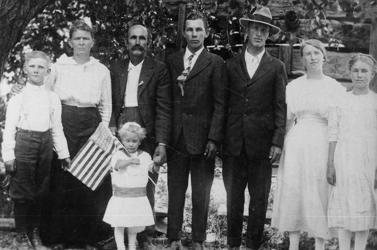 John R. Crawford family assembled outdoors during the WWI era (1918). Left to right: Norman Pierce, Elisa Crawford, John R. Crawford, Verda Crawford, Davis Peterson in front holding US Flag, Newell Knight, William, Susan Crawford Ruesch, Lucy Crawford Schaffer. Squire was in the service and is not pictured.