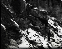 Man standing beneath halfpipe culvert on steep slope, snow on ground.