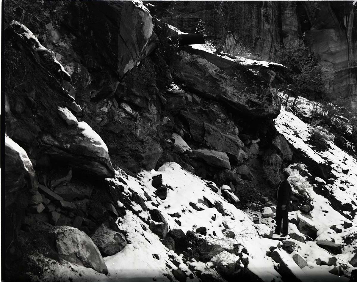 Man standing beneath halfpipe culvert on steep slope, snow on ground.