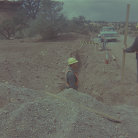 Man standing in trench off side of road while other men construct and repair roads.