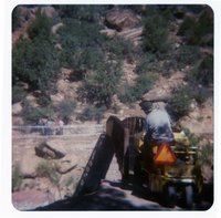 Man steering sweeper equipment onto the new Grotto footbridge after its placement on stone abutments. Men working on trail in background.