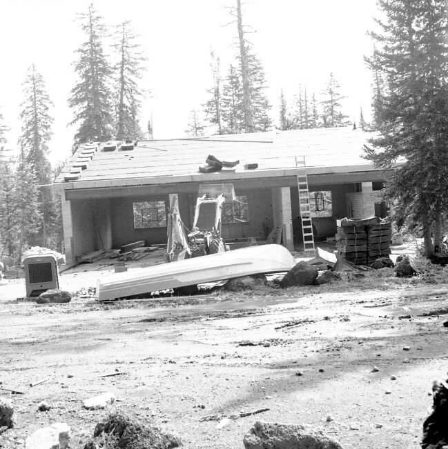 Utility and maintenance building under construction with stacks of shake shingles on roof. Supplies and equipment alongside downturned boat in front of building.