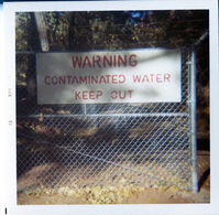 Sign reading 'Warning contaminated water keep out' near a pond of contaminated water or waste.