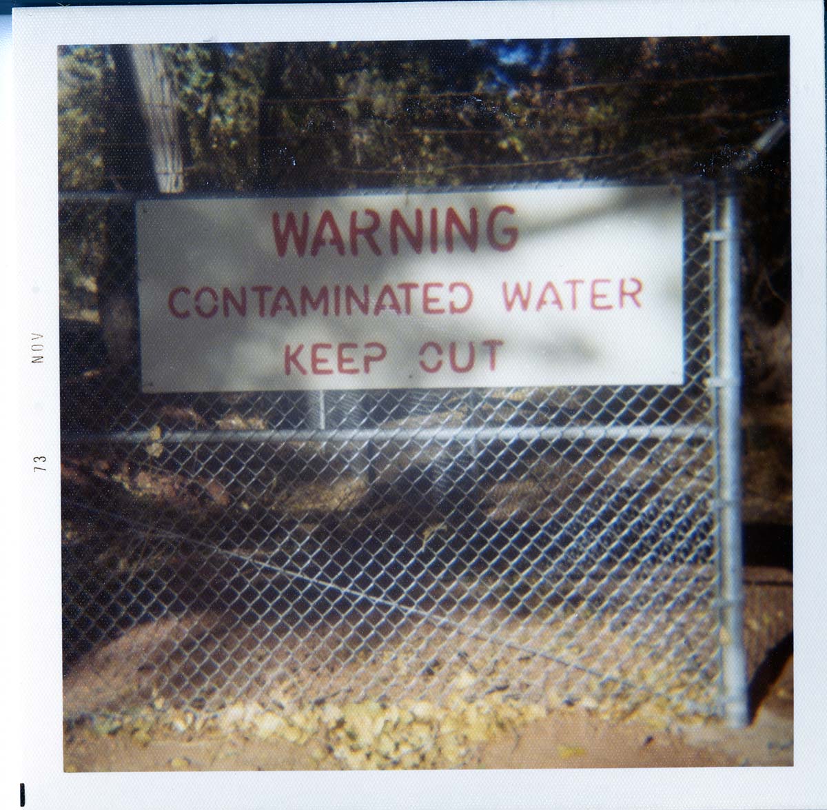 Sign reading 'Warning contaminated water keep out' near a pond of contaminated water or waste.