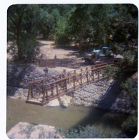 Men on new Grotto footbridge as it is lowered into place by pulley system, crane and backhoe anchoring one end of bridge. Note riverbank revetments.