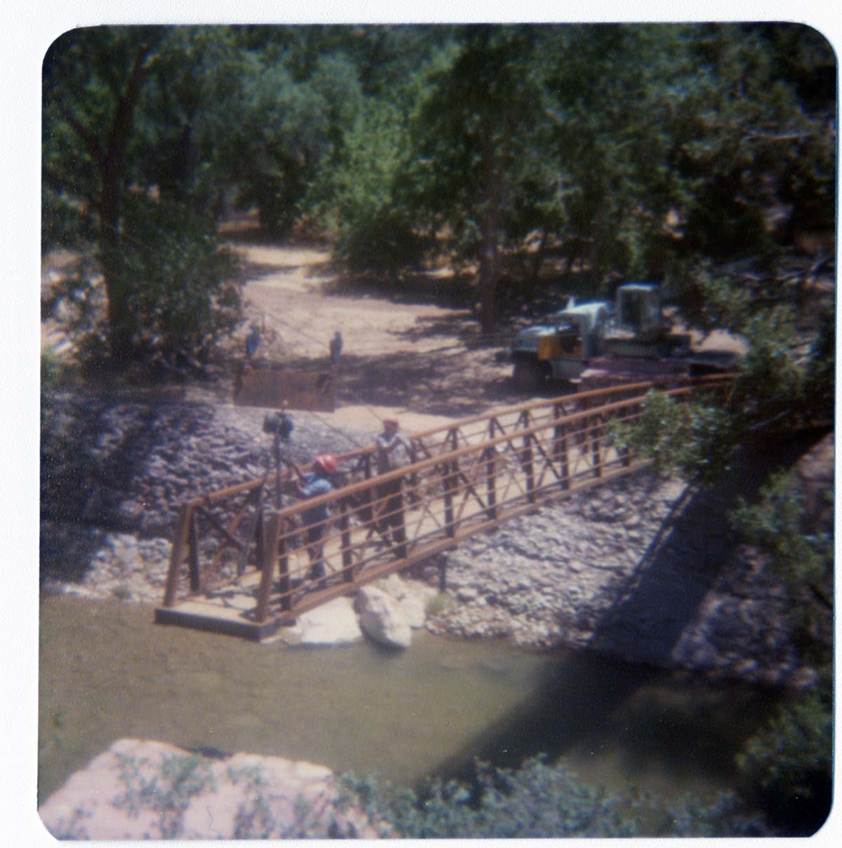 Men on new Grotto footbridge as it is lowered into place by pulley system, crane and backhoe anchoring one end of bridge. Note riverbank revetments.
