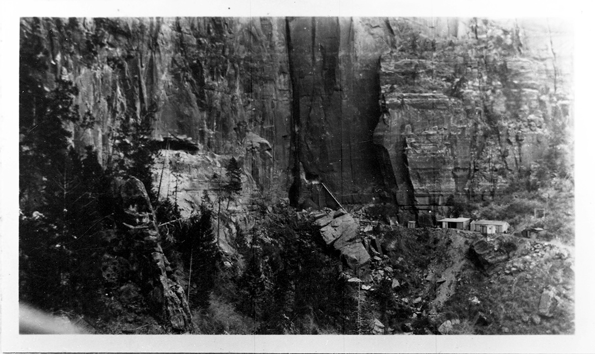 Galleries 1 & 6 under construction, stairway suspended on iron bars protruding from cliff shows to right of gallery #6 blacksmith, compressor, and transformer houses on trail around talus slope.