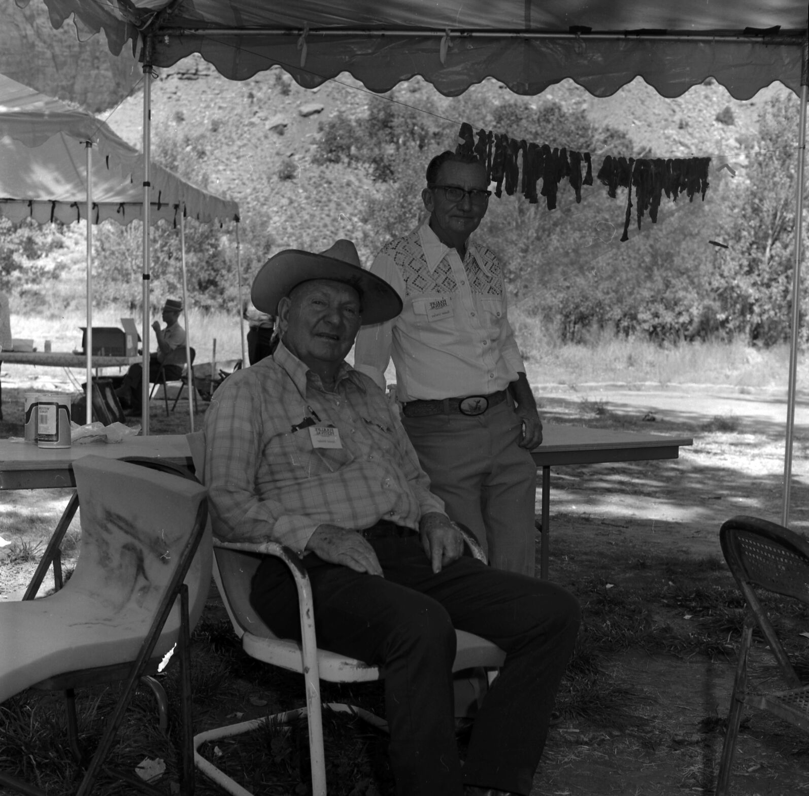 Dan Winder (seated) and Rueben Crawford demonstrating the making of jerky at first annual Folklife Festival, Zion National Park Nature Center, September 1977. Jerky hanging on cord in background.