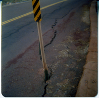 Crack in road starting at the base of a black and yellow striped construction sign.