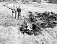 Two men watching water level in trench opened for water exploration, nearby large dirt pile. Water pump and gauge set up in trench.