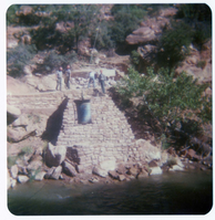 Four men standing on stone abutment for new Grotto footbridge as supplies are sent by pulley across Virgin River.