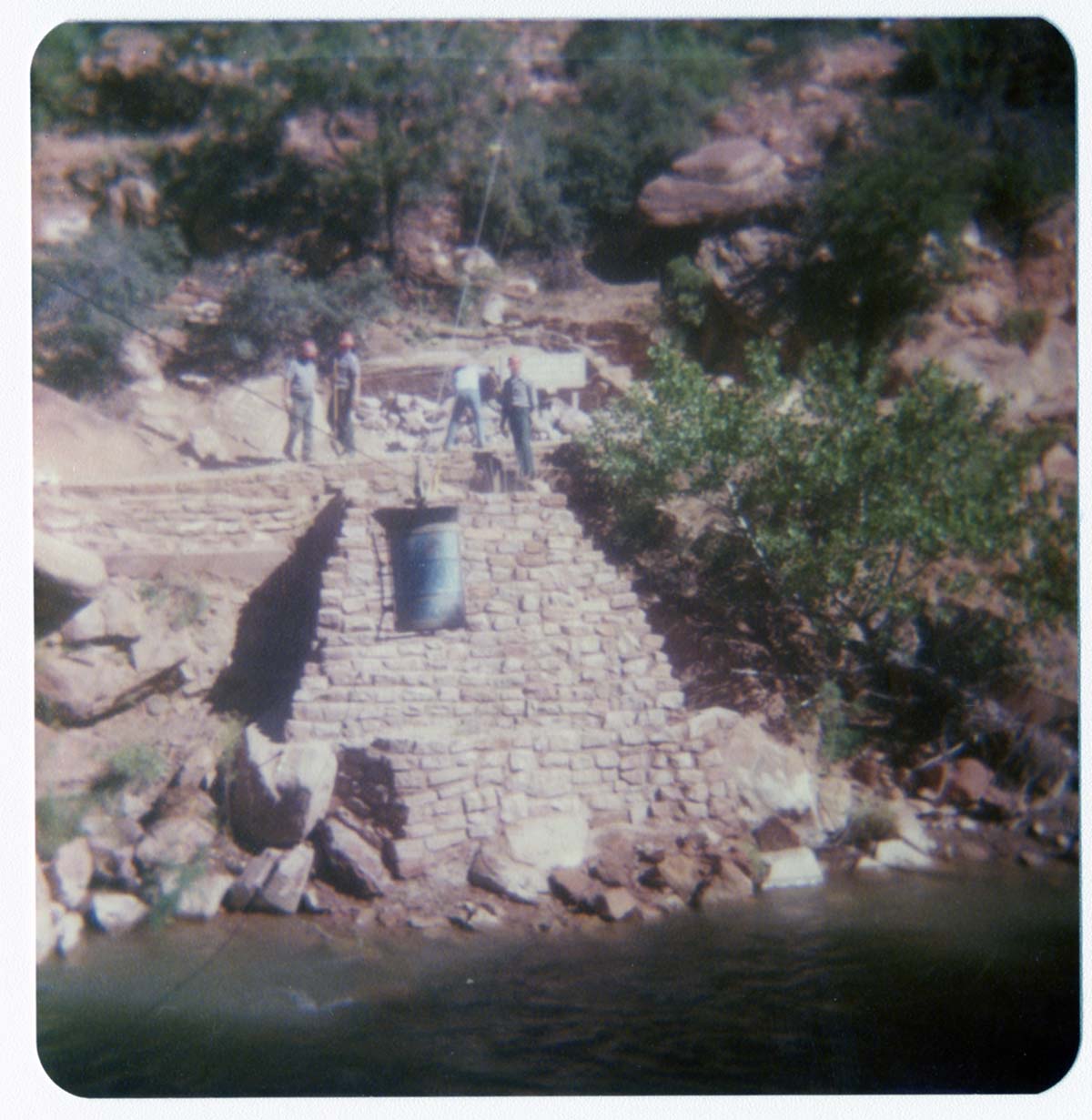 Four men standing on stone abutment for new Grotto footbridge as supplies are sent by pulley across Virgin River.