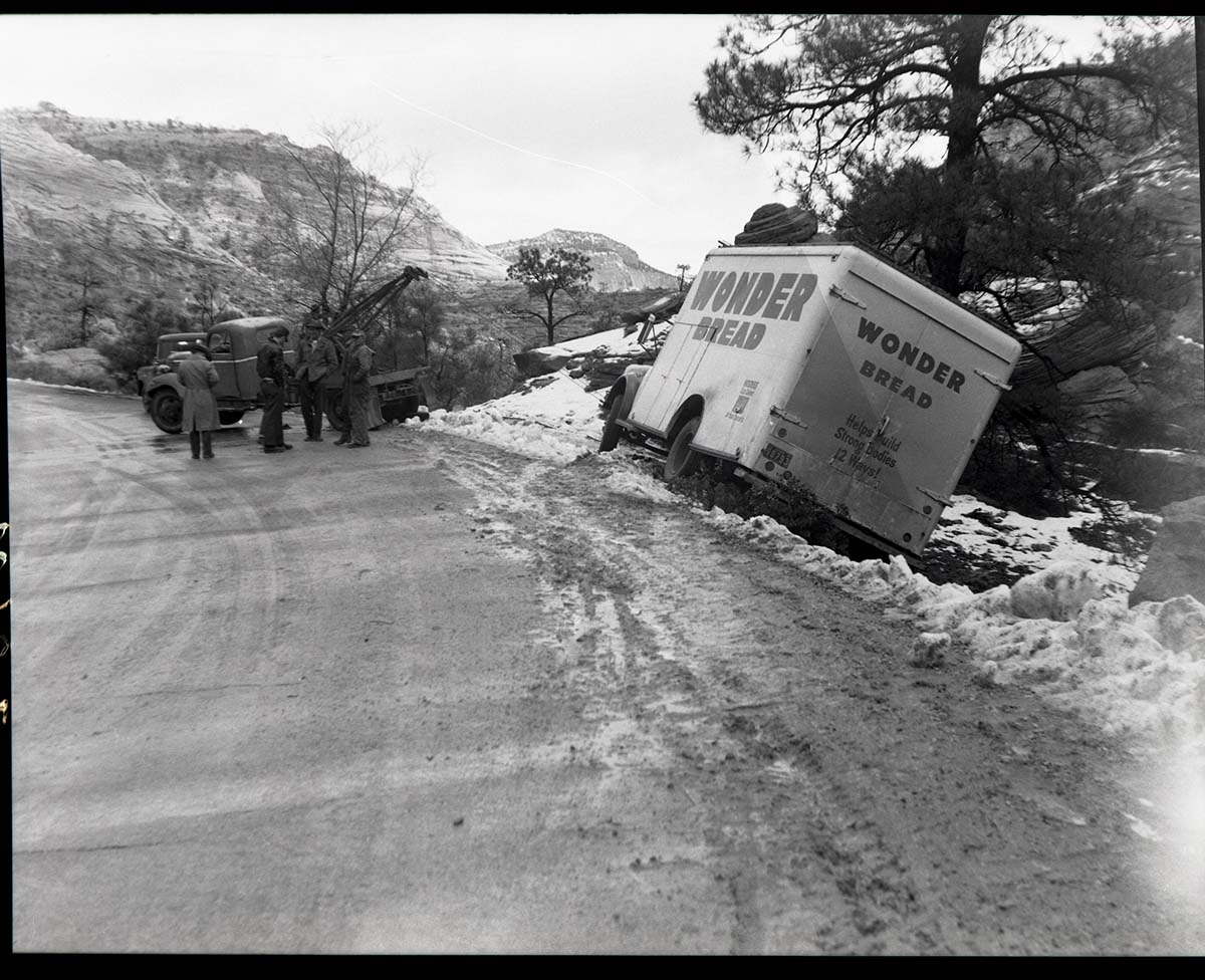 Accident involving a Wonder Bread truck which slid off road east of small tunnel because of icy road and too much speed, saved by large pine tree. Wrecker and several people assisting.