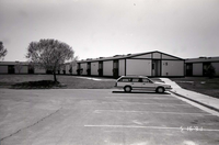 Barracks and parking area with man walking on sidewalk. Building number 5. [Image possibly for comparative housing study]